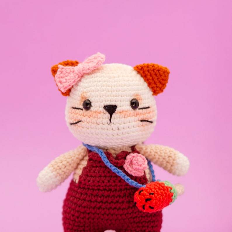 May include: A crocheted cat toy with a pink bow, white fur, red overalls, and a blue strap with a pink flower and a red strawberry. The toy is against a pink background.