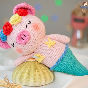 May include: A pink crocheted pig wearing a mermaid tail with blue and green accents. The pig has a flower crown with red, yellow, and blue flowers. The pig is lying on a yellow and white sea urchin.
