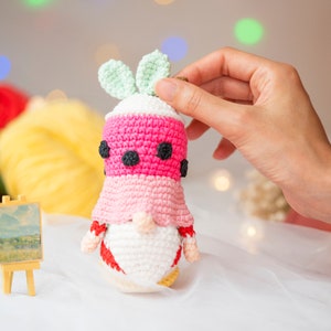 May include: A crocheted gnome doll wearing a pink and white watermelon costume with black seed accents. The gnome has a white beard and a green leaf on top of its head.