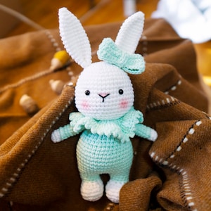 May include: A crocheted bunny rabbit toy with white fur, a light blue and green outfit, and a light blue bow. The toy is sitting on a brown blanket.