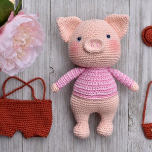 May include: A crocheted pink pig wearing a pink and white striped shirt. The pig is holding a small brown crocheted purse. There is a brown crocheted hat and a brown crocheted pair of shorts in the image.