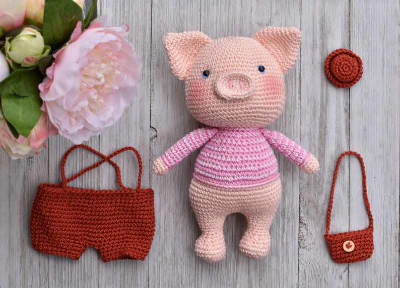 May include: A crocheted pink pig wearing a pink and white striped shirt. The pig is holding a small brown crocheted purse. There is a brown crocheted hat and a brown crocheted pair of shorts in the image.