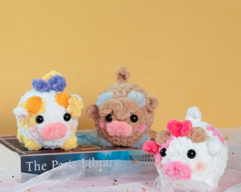 Puede incluir: Tres peluches de vaca de ganchillo hechos a mano en blanco, marrón y rosa. Cada peluche tiene un diseño único, incluyendo lazos y detalles florales. Los peluches están sobre un libro y una tela de tul rosa.
