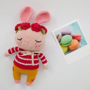 May include: A pink crocheted bunny with red and green flower accents, wearing a red and white striped shirt and yellow pants.