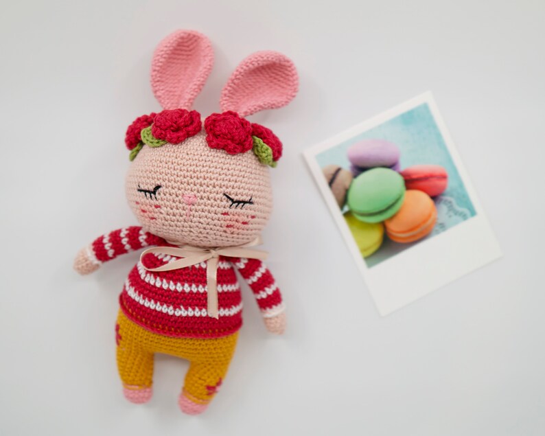 May include: A pink crocheted bunny with red and green flower accents, wearing a red and white striped shirt and yellow pants.