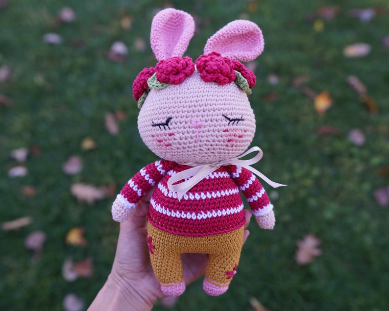 May include: A pink crocheted bunny with a red and white striped shirt, brown pants, and a flower crown. The bunny has closed eyes and a sweet expression.