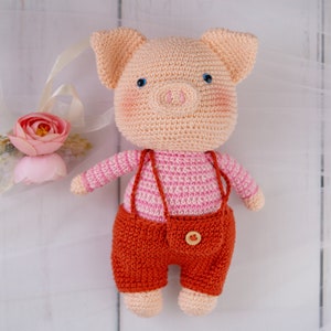 May include: A crocheted pig toy wearing a pink and white striped shirt and orange overalls. The pig has a pink snout and blue eyes.