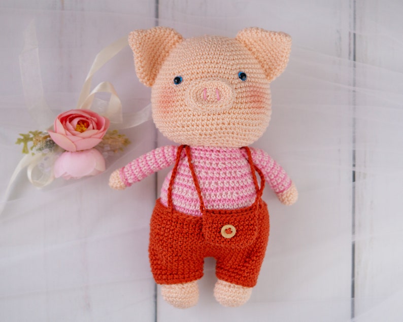 May include: A crocheted pig toy wearing a pink and white striped shirt and orange overalls. The pig has a pink snout and blue eyes.
