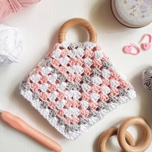 May include: A crocheted baby teether with a wooden ring, featuring a pink, white, and gray color scheme. The teether is made with a granny square pattern.