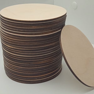 May include: A stack of round, wooden craft blanks. The blanks are light brown and have a smooth, unfinished surface. They are stacked neatly, with the top blank slightly offset from the others. A single blank is lying on its side next to the stack.