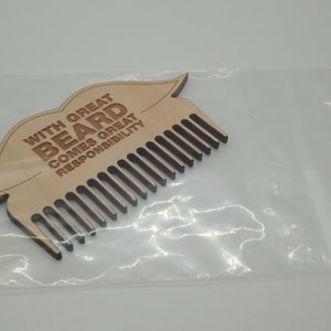 Op de afbeelding: Een houten baardkam in de vorm van een mond met de tekst "With great beard comes great responsibility" erop gegraveerd. De kam is verpakt in een doorzichtige plastic zak.