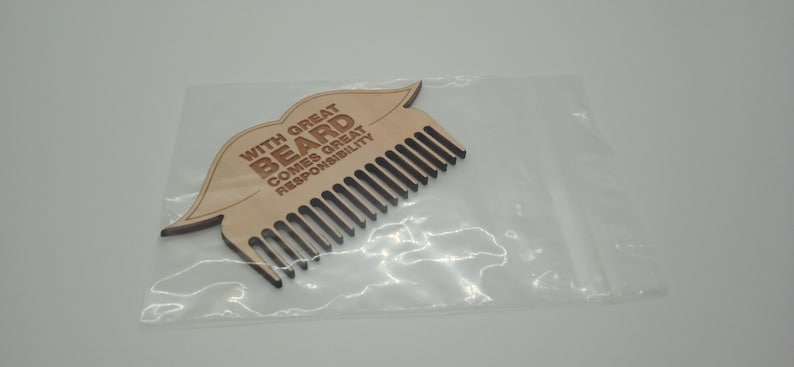 Op de afbeelding: Een houten baardkam in de vorm van een mond met de tekst "With great beard comes great responsibility" erop gegraveerd. De kam is verpakt in een doorzichtige plastic zak.