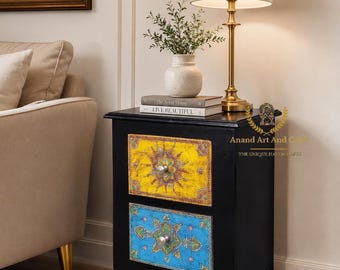 May include: A black wooden nightstand featuring two drawers with hand-painted yellow and blue mandala-style patterns, each equipped with a single silver knob. The text Anand Art And Craft The Unique Handicraft is visible on the side of the unit.