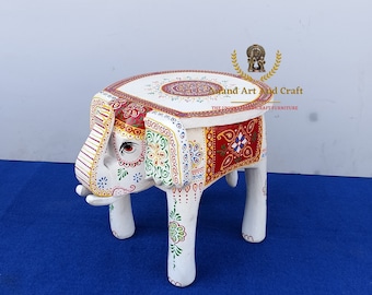 May include: A white wooden stool shaped like an elephant with hand-painted floral and geometric patterns in red, green, yellow, and blue. The top features a circular mandala design, and the text Anand Art And Craft is visible above the stool.