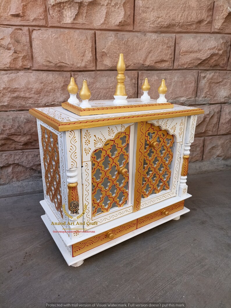 Op de afbeelding: Een witte houten kast met goudkleurige accenten, voorzien van twee deuren met ingewikkelde rasteruitsnijdingen, een bovenplatform met vijf decoratieve knoppen en een onderste lade. De tekst Anand Art And Craft staat op de voorzijde.