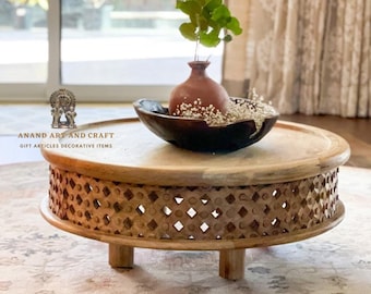May include: A round wooden coffee table with a carved lattice pattern on the side and short legs. On top sits a dark bowl containing a small terracotta vase with green leaves and white flowers. Text reads ANAND ART AND CRAFT GIFT ARTICLES DECORATIVE ITEMS.