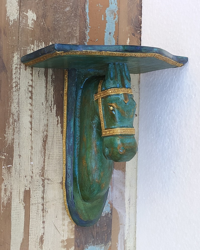May include: A wall-mounted shelf featuring a sculpted horse head support. The horse is finished in a distressed teal and green patina with gold-toned trim along the bridle and the edges of the shelf. The shelf has a flat top surface.
