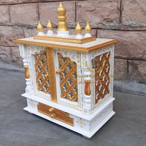 Op de afbeelding: Een witte houten huistempel met goudkleurige accenten, ingewikkelde latwerk deurpanelen en een lade. De bovenkant is versierd met vijf gouden sierspitsen. De tekst Anand Art And Craft is zichtbaar op het voorpaneel van de structuur.