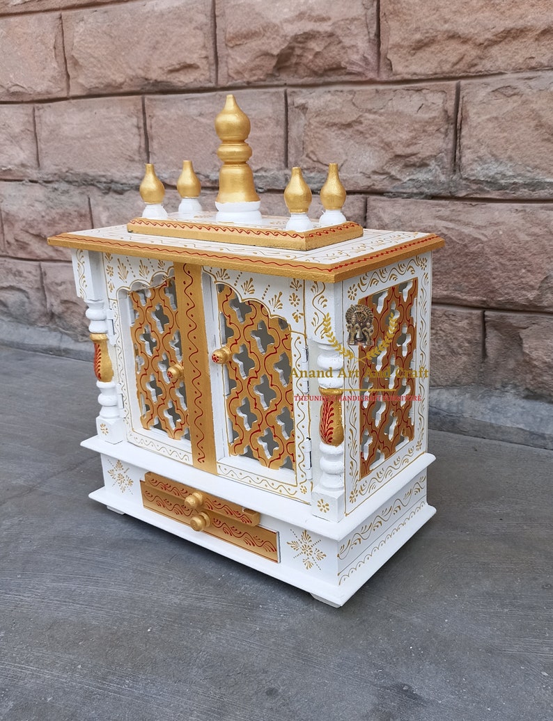 Op de afbeelding: Een witte houten huistempel met goudkleurige accenten, ingewikkelde latwerk deurpanelen en een lade. De bovenkant is versierd met vijf gouden sierspitsen. De tekst Anand Art And Craft is zichtbaar op het voorpaneel van de structuur.