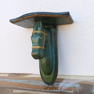 May include: A wall-mounted shelf featuring a horse head sculpture as the support bracket. The piece is finished in a distressed teal and green patina with ornate gold-coloured trim detailing along the edges of the shelf and the horse's bridle.