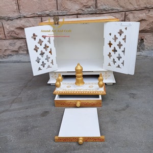 Op de afbeelding: Een witte houten huistempel met gouden accenten, voorzien van twee open deuren met ingewikkelde uitgesneden patronen, een uitschuifbare lade en een afneembaar platform. De tekst Anand Art And Craft is zichtbaar op het achterpaneel binnenin.