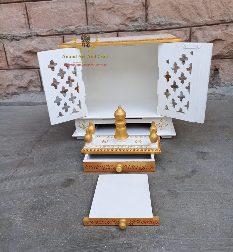 Op de afbeelding: Een witte houten huistempel met gouden accenten, voorzien van twee open deuren met ingewikkelde uitgesneden patronen, een uitschuifbare lade en een afneembaar platform. De tekst Anand Art And Craft is zichtbaar op het achterpaneel binnenin.