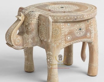 May include: A light brown wooden stool shaped like an elephant with a flat circular top. The surface features intricate white painted floral patterns and small embedded circular mirrors. The text Anand Art And Craft is visible on the side.