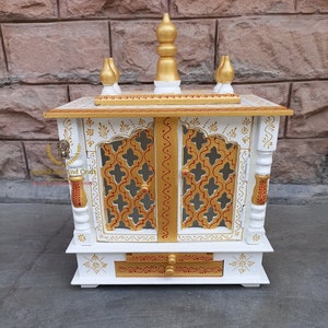 Op de afbeelding: Een witte houten huistempel met goudkleurige accenten, ingewikkelde rasterdeuren en een centrale lade. De structuur is bekroond met een gouden topstuk en vier kleinere decoratieve spitsen. De tekst Anand Art And Craft is zichtbaar.