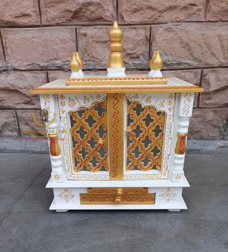 Op de afbeelding: Een witte houten huistempel met goudkleurige accenten, ingewikkelde rasterdeuren en een centrale lade. De structuur is bekroond met een gouden topstuk en vier kleinere decoratieve spitsen. De tekst Anand Art And Craft is zichtbaar.