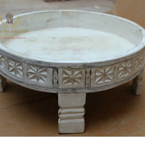 May include: A round, white-washed wooden tray with four short, carved legs. The exterior side features a repeating pattern of carved star-like geometric shapes. The text 'Anand Art And Craft THE UNIQUE HANDICRAFT FURNITURE' is visible in the upper left.