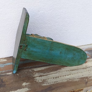 May include: A teal-coloured wooden wall shelf with a distressed finish and a flat, rectangular back panel. The shelf features a rounded front edge and is accented with a thin, gold-coloured decorative trim along the top perimeter of the horizontal surface.