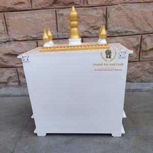 Op de afbeelding: Een witte houten huistempel met een goudkleurige bovenkant en vijf decoratieve ornamenten. Het bovenoppervlak heeft gouden details. De voorzijde toont de tekst Anand Art And Craft met een logo. Twee metalen ringen zijn aan de zijkanten bevestigd.