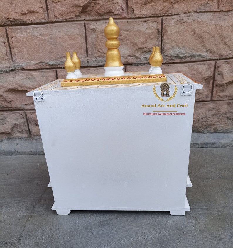 Op de afbeelding: Een witte houten huistempel met een goudkleurige bovenkant en vijf decoratieve ornamenten. Het bovenoppervlak heeft gouden details. De voorzijde toont de tekst Anand Art And Craft met een logo. Twee metalen ringen zijn aan de zijkanten bevestigd.