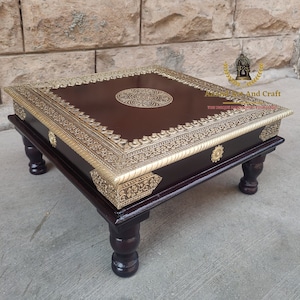 Könnte beinhalten: Ein quadratischer, dunkelbrauner Holztisch mit niedrigen Beinen, verziert mit goldfarbenen Metallkanten und einem kreisförmigen Motiv in der Mitte. Der Tisch steht auf vier gedrechselten Beinen und trägt den Text Anand Art And Craft.