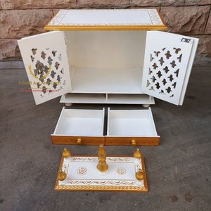 Op de afbeelding: Een witte houten kast met gouden randen, voorzien van twee deuren met complexe uitgesneden patronen, twee uitschuifbare lades en een aparte rechthoekige basis met vier kleine goudkleurige zuilen en een centrale gouden knop.