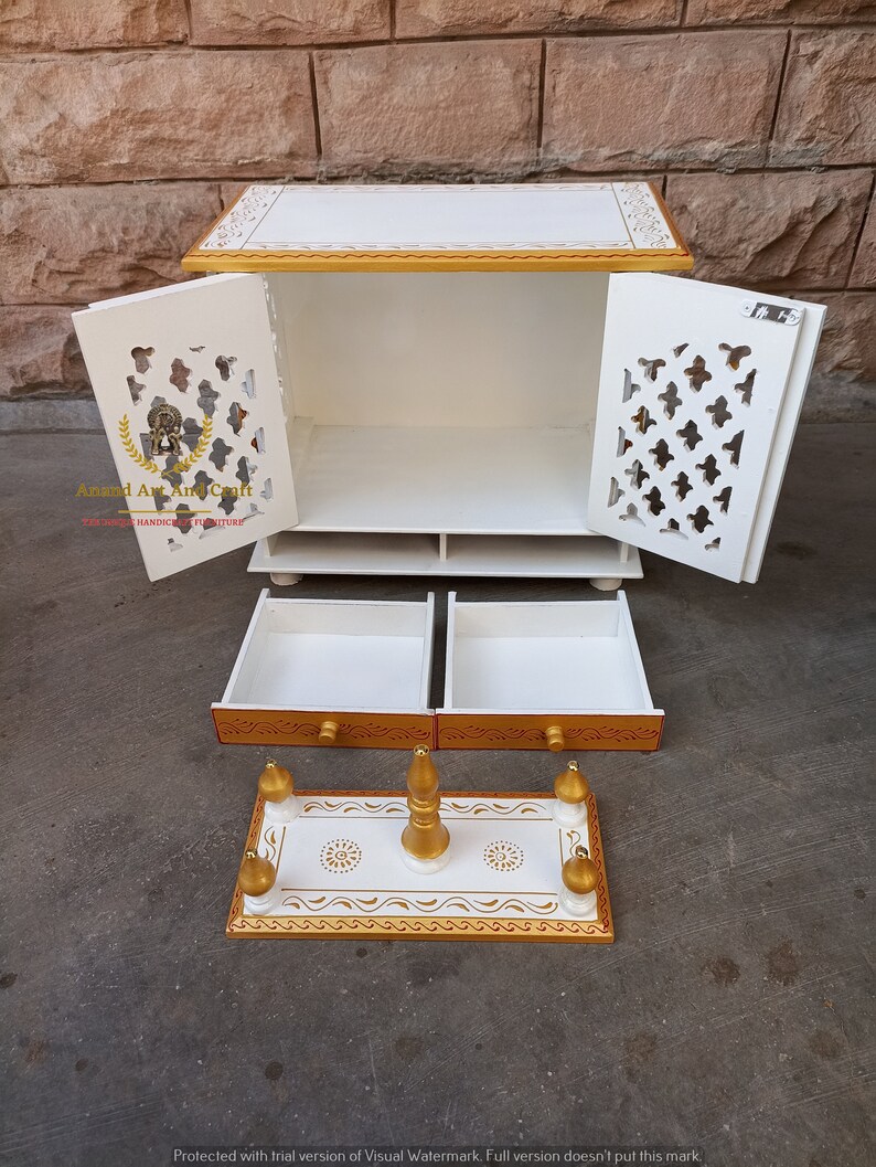 Op de afbeelding: Een witte houten kast met gouden randen, voorzien van twee deuren met complexe uitgesneden patronen, twee uitschuifbare lades en een aparte rechthoekige basis met vier kleine goudkleurige zuilen en een centrale gouden knop.