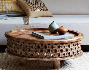 May include: A round wooden coffee table with a carved lattice pattern along the side, featuring a natural brown finish. On the tabletop sits a light gray ceramic vessel, a small brown wooden lidded container, and a book titled BRANCUSI.