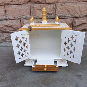 Op de afbeelding: Een witte houten huistempel met twee deuren met ingewikkelde rasteruitsnijdingen, een uitschuifbare lade met goudkleurige accenten en een bovenste laag versierd met vijf gouden sierspitsen. De tekst Anand Art And Craft is zichtbaar op de linker deur.