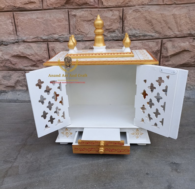 Op de afbeelding: Een witte houten huistempel met twee deuren met ingewikkelde rasteruitsnijdingen, een uitschuifbare lade met goudkleurige accenten en een bovenste laag versierd met vijf gouden sierspitsen. De tekst Anand Art And Craft is zichtbaar op de linker deur.