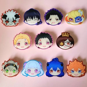 May include: Twelve colourful anime character face magnets. Each magnet features a unique character design with distinct hairstyles, eye colours, and facial expressions. The magnets are arranged on a light pink surface.
