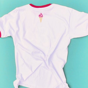 May include: White t-shirt with red trim on the collar and sleeves. A pink and white ice cream cone graphic is printed on the back. The shirt is made of a soft, lightweight material.