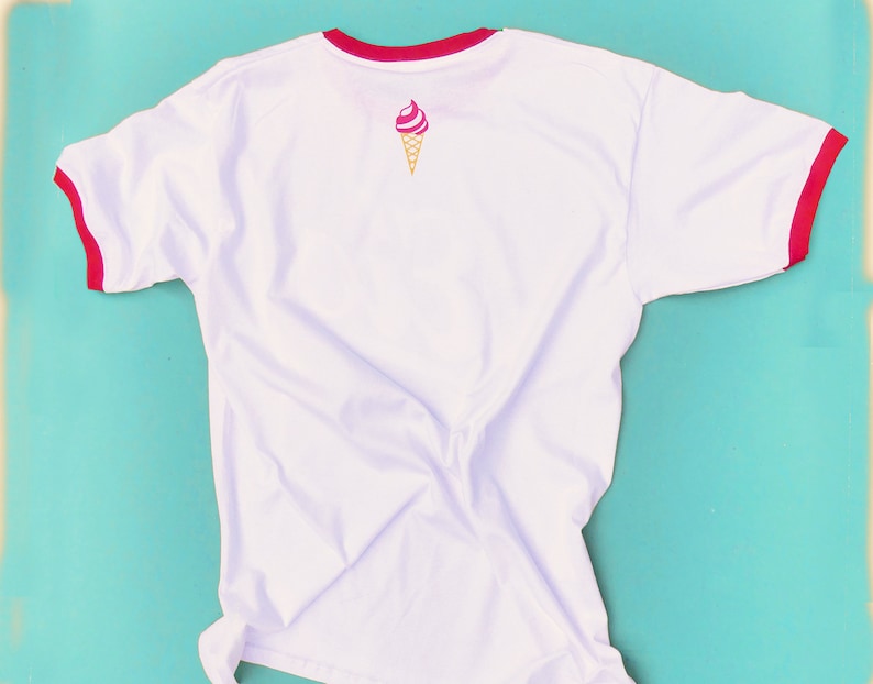 May include: White t-shirt with red trim on the collar and sleeves. A pink and white ice cream cone graphic is printed on the back. The shirt is made of a soft, lightweight material.