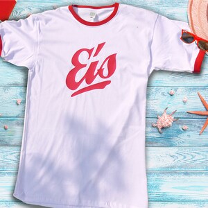 May include: White t-shirt with red trim and the word "Eis" in red script. The shirt is displayed on a blue wooden surface with beach-themed accessories, including a starfish, seashells, and sunglasses.