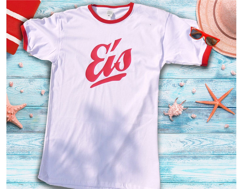 May include: White t-shirt with red trim and the word "Eis" in red script. The shirt is displayed on a blue wooden surface with beach-themed accessories, including a starfish, seashells, and sunglasses.