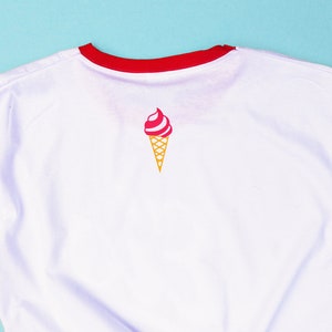 May include: White t-shirt with a red collar and a small ice cream cone graphic. The ice cream cone has a pink and white swirl with a yellow cone. The shirt is made of a soft material.