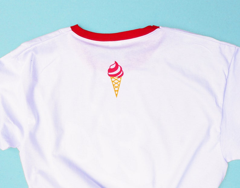 May include: White t-shirt with a red collar and a small ice cream cone graphic. The ice cream cone has a pink and white swirl with a yellow cone. The shirt is made of a soft material.