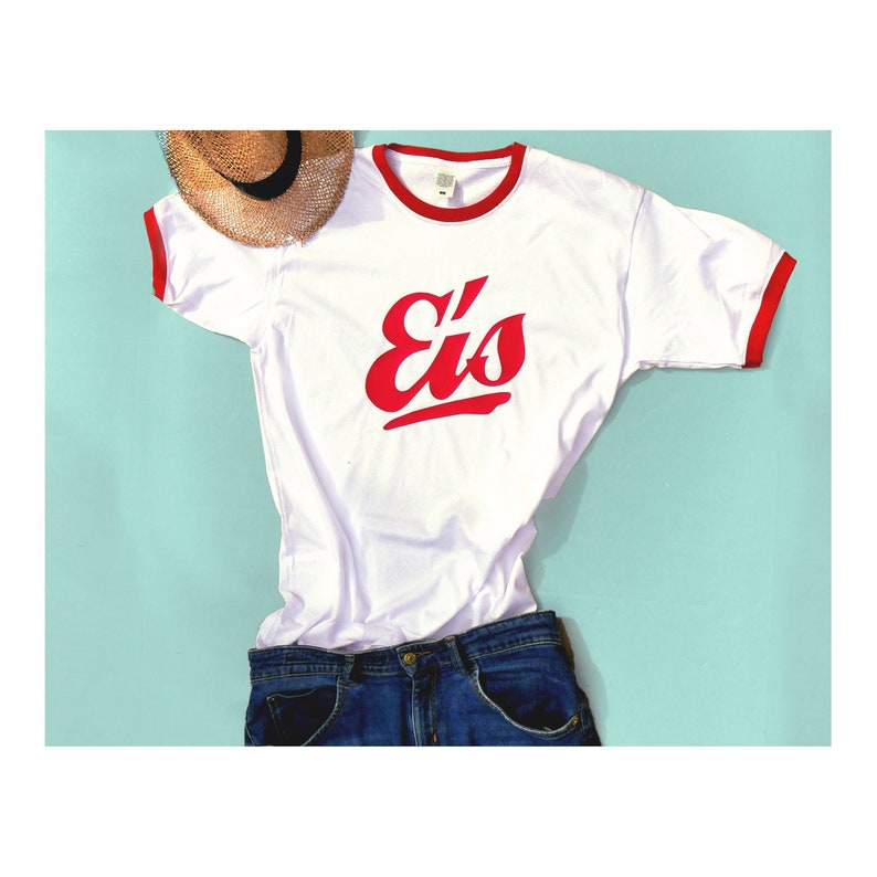 Könnte beinhalten: Weißes T-Shirt mit roten Rippbündchen und einem roten "Eis"-Logo auf der Vorderseite. Das Shirt liegt auf einem hellblauen Untergrund mit einem Strohhut.