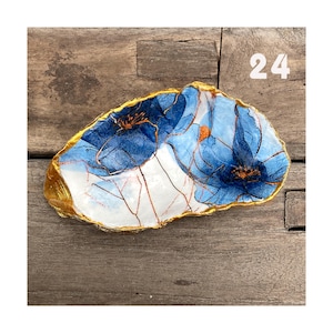 Könnte beinhalten: Eine unregelmäßig geformte dekorative Schale mit goldenem Rand und floralem Design. Die Schale zeigt blaue Blumen mit goldenen Akzenten auf weißem Hintergrund. Die Nummer "24" ist oben rechts sichtbar.
