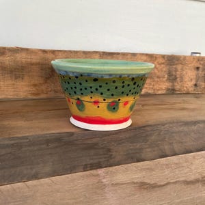 May include: A hand-painted ceramic bowl with a unique design. The bowl features a green rim, a band of green with black dots, a yellow band with red accents, and a red base. The bowl is set on a wooden surface.