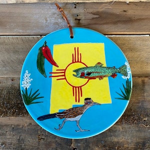 May include: A blue ceramic plate with a yellow background featuring a stylized image of the New Mexico state flag, a red chili pepper, a green chili pepper, a trout, and a roadrunner.